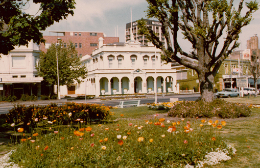 Historical image in colour showing a side view of the town hall and the park in in front, with people in the street