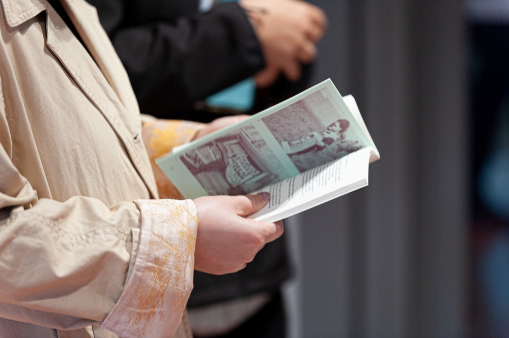 A person in a beige coat holding an open booklet with images and text, appearing focused. Another person is blurred in the background. The setting is calm and introspective.