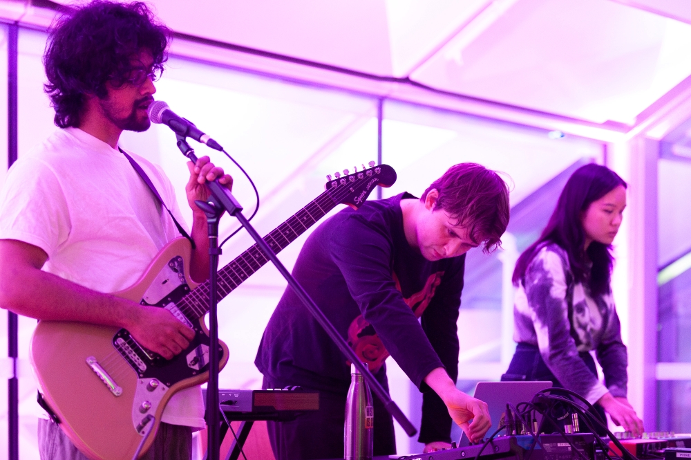 A three-person band performs in a room bathed in purple light. The guitarist sings into a mic, while two others use electronic equipment, creating a focused atmosphere.