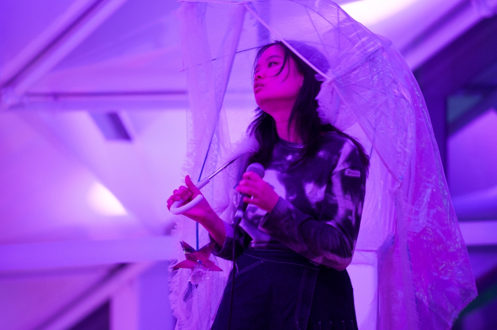 A person stands holding a transparent umbrella and a microphone under purple lighting, conveying an artistic and contemplative mood.