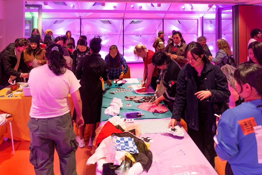 A group of people gather around tables covered with colorful fabrics in a well-lit room. The atmosphere is lively and collaborative, with a purple-lit backdrop.