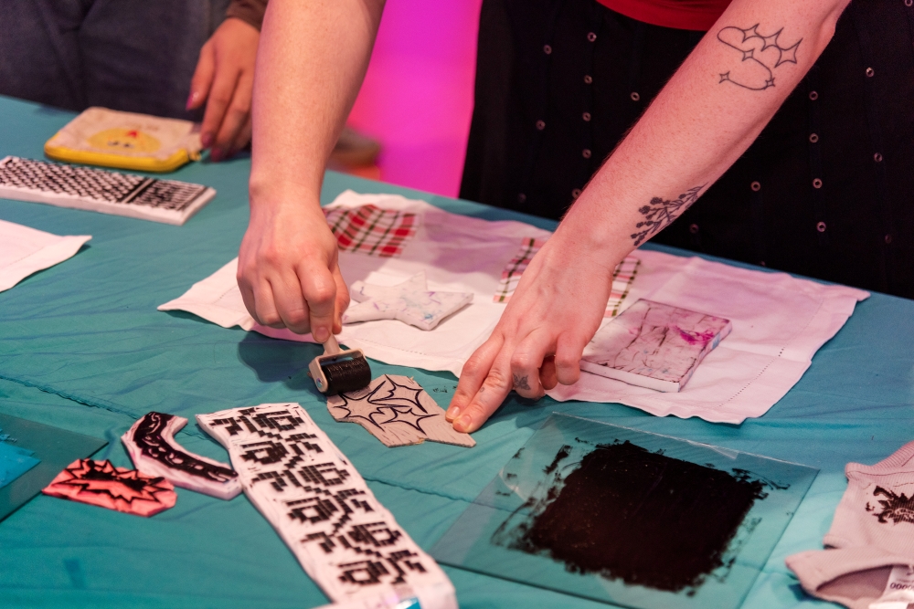 Two hands engaged in a fabric printing activity. One hand holds a roller with black ink, while the other points to a fabric stencil on a teal table. Art supplies and patterned fabrics surround the scene, conveying creativity and focus.