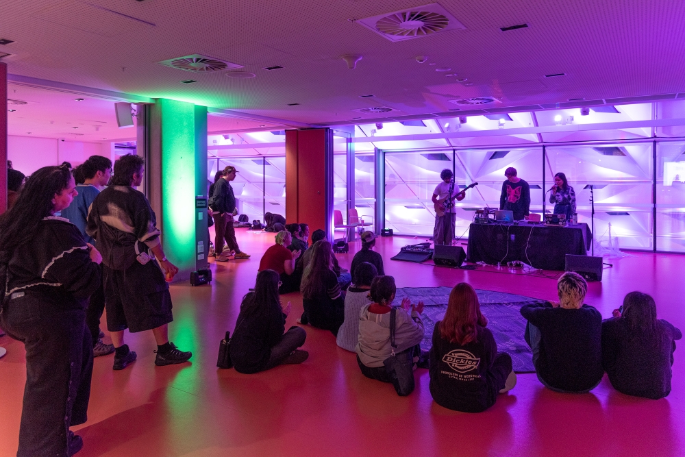 Indoor music performance with a band playing on stage. Seated audience watches them in a room lit by purple and green lights, creating a vibrant atmosphere.
