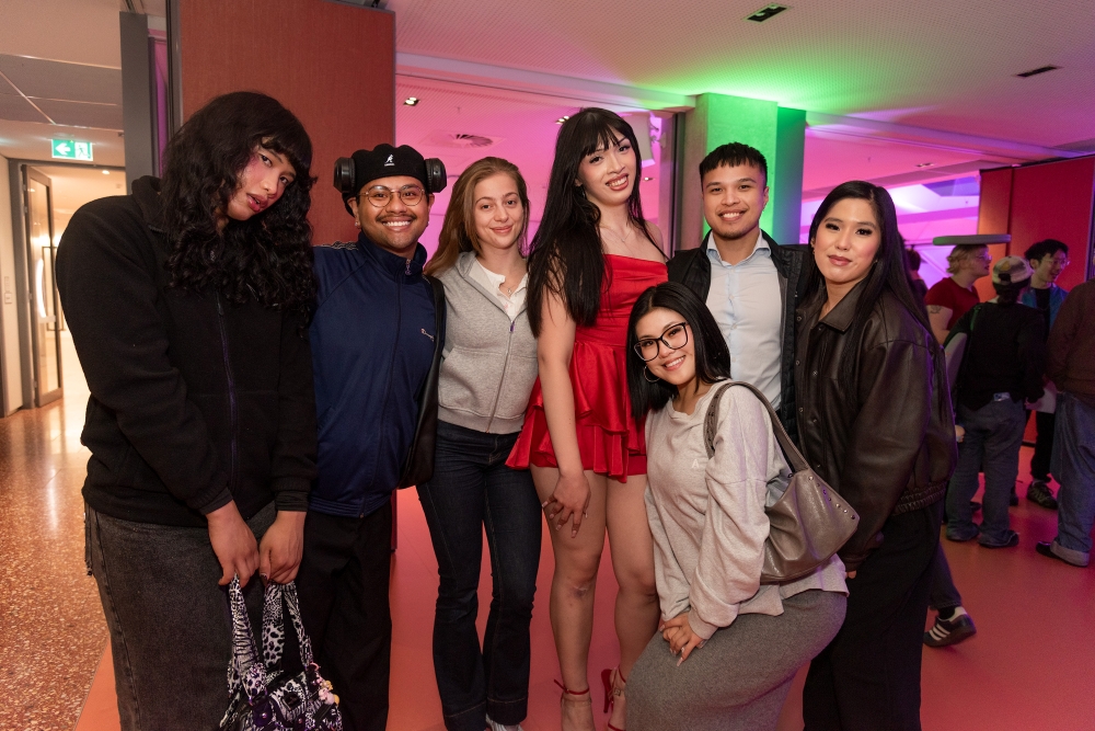 A group of seven people smiling and posing in a warmly-lit room with colorful lighting. They appear happy and are dressed in various casual and stylish outfits.