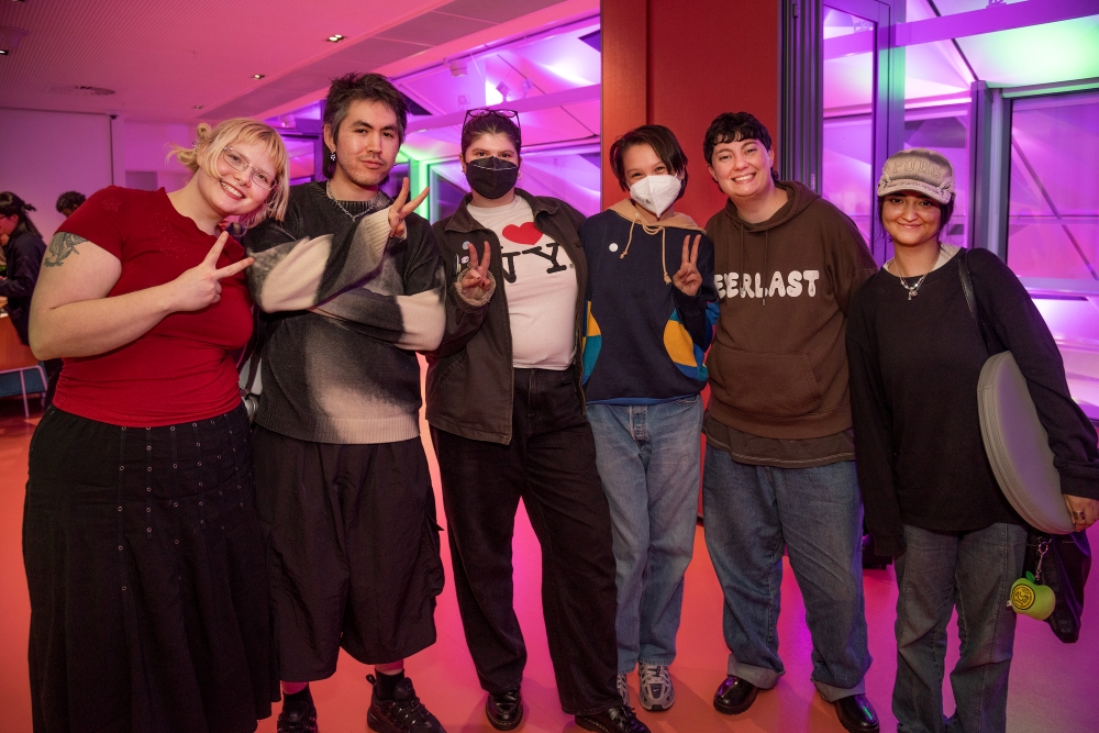 A diverse group of six people pose indoors, smiling and making peace signs. They wear casual, eclectic outfits. The background is lit with purple and green lights, creating a vibrant, joyful atmosphere.