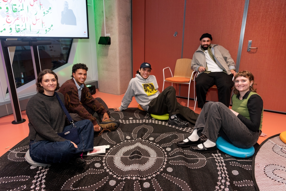 Five people sit casually on a patterned rug in a modern room, smiling and relaxed. A screen displays colorful text and an image in the background.