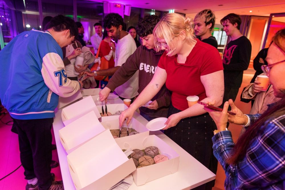 A diverse group of people at a gathering in a room with purple and orange lighting. They're joyfully serving themselves from boxes of mochi at a table.
