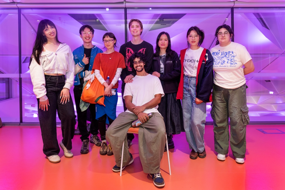 A group of eight friends poses cheerfully on a vibrant orange floor with a purple-lit glass wall backdrop. One person sits on a chair smiling. Casual attire and a lively atmosphere convey a sense of camaraderie and fun.