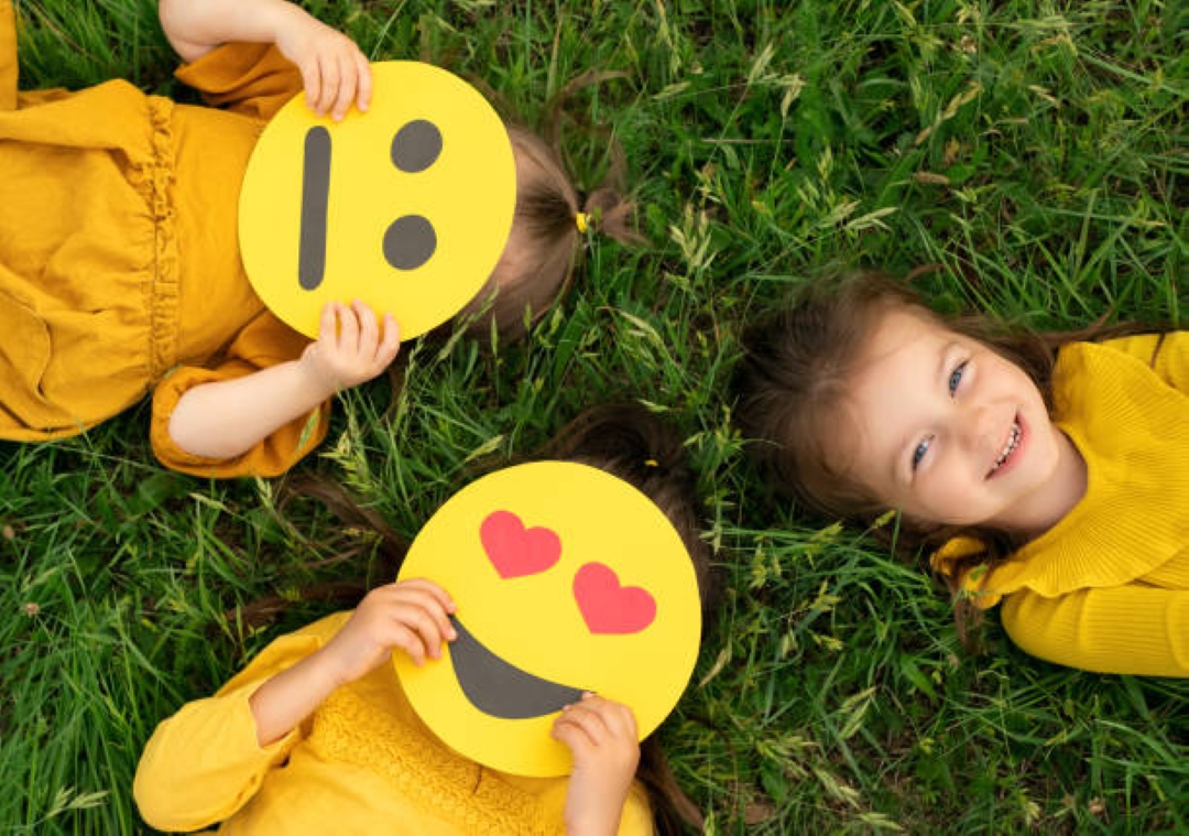 Three children lie on grass, two holding emoji faces with different expressions. They wear yellow dresses, creating a playful and cheerful scene.