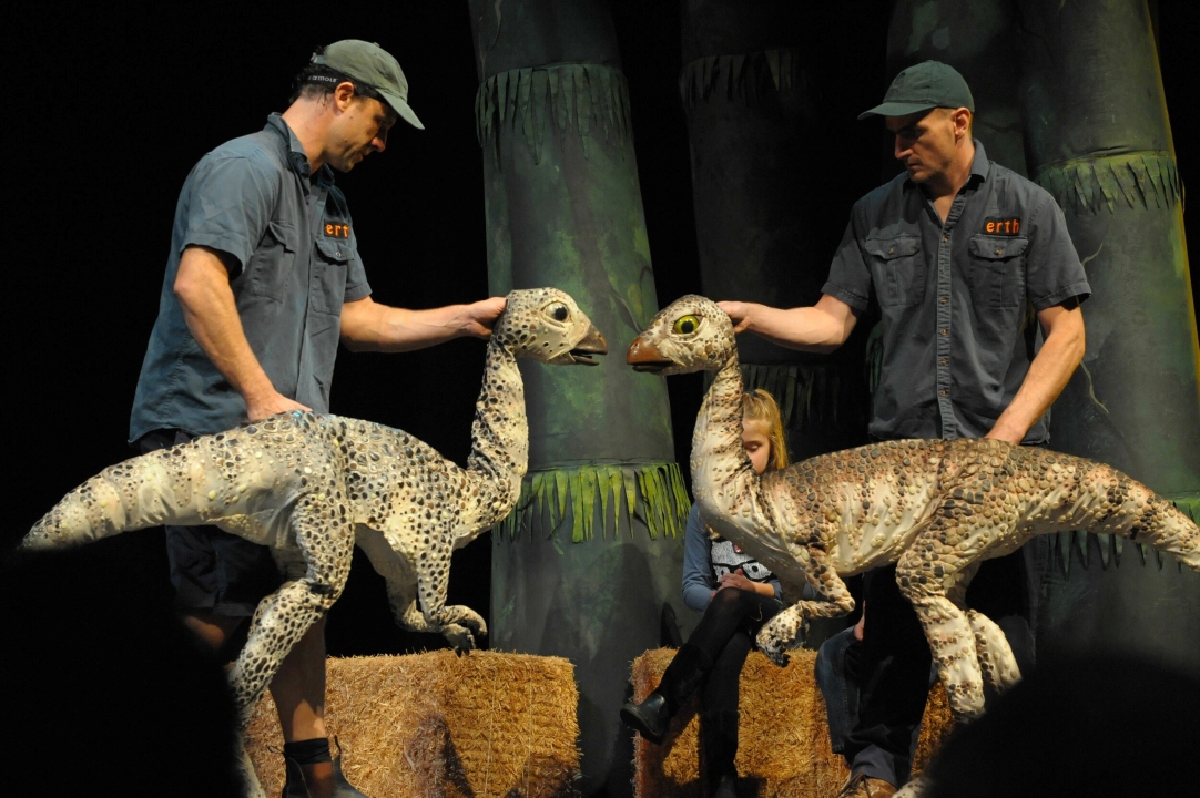 Two performers manipulate realistic dinosaur puppets on stage, dressed in casual blue uniforms. A child sits nearby, captivated. The scene feels lively and engaging.