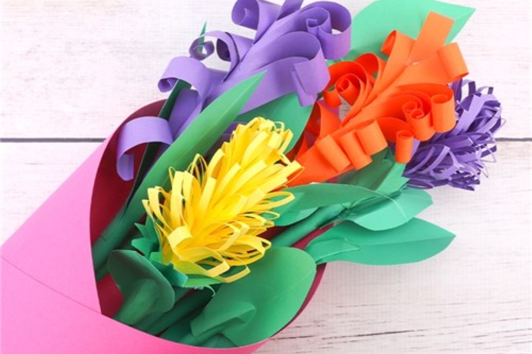 A vibrant bouquet of paper flowers on a wooden surface. The flowers, in purple, orange, and yellow, have curly petals and are wrapped in pink paper.