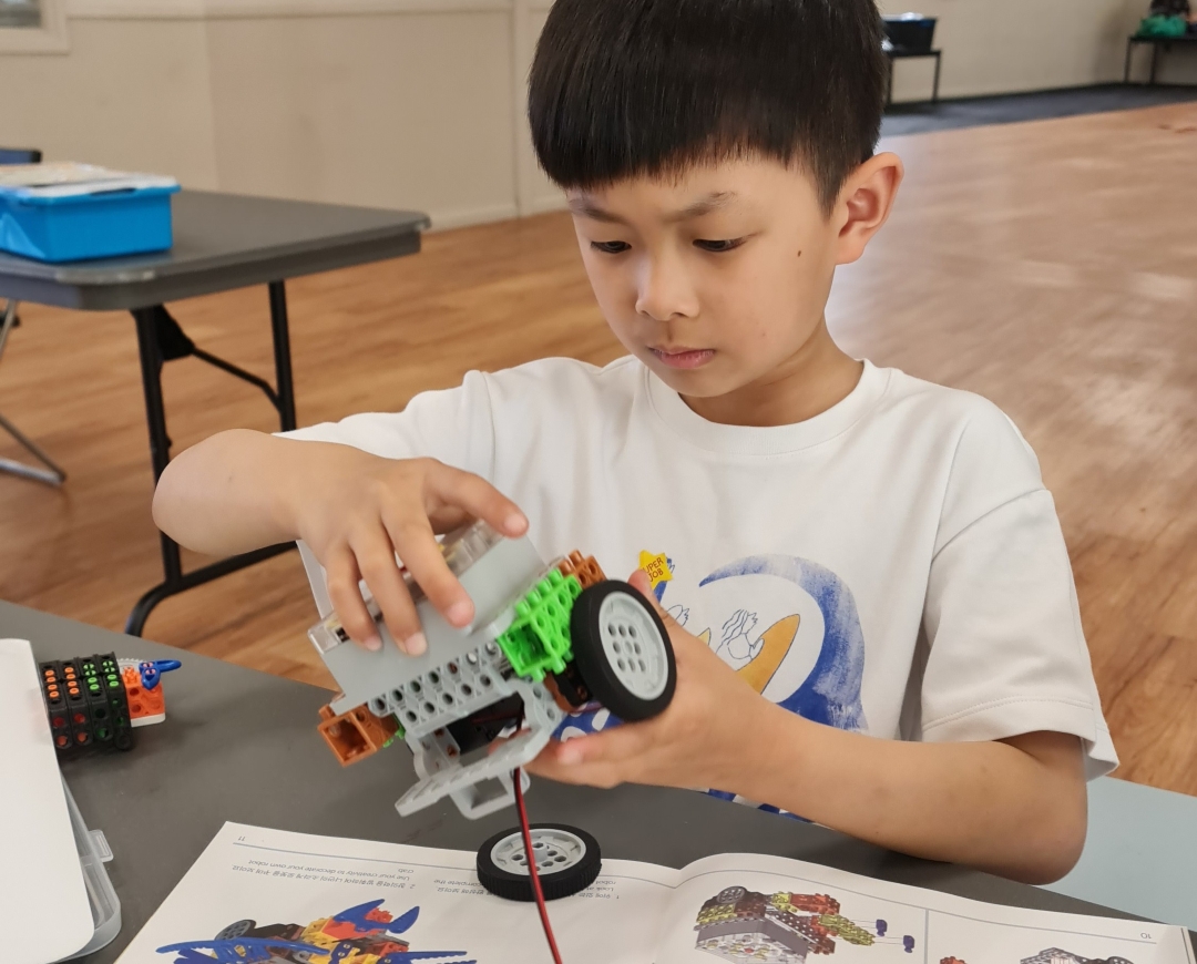 A young boy focuses intently on assembling a LEGO robot vehicle using a kit, with various colored blocks and wheels. The setting is a classroom.