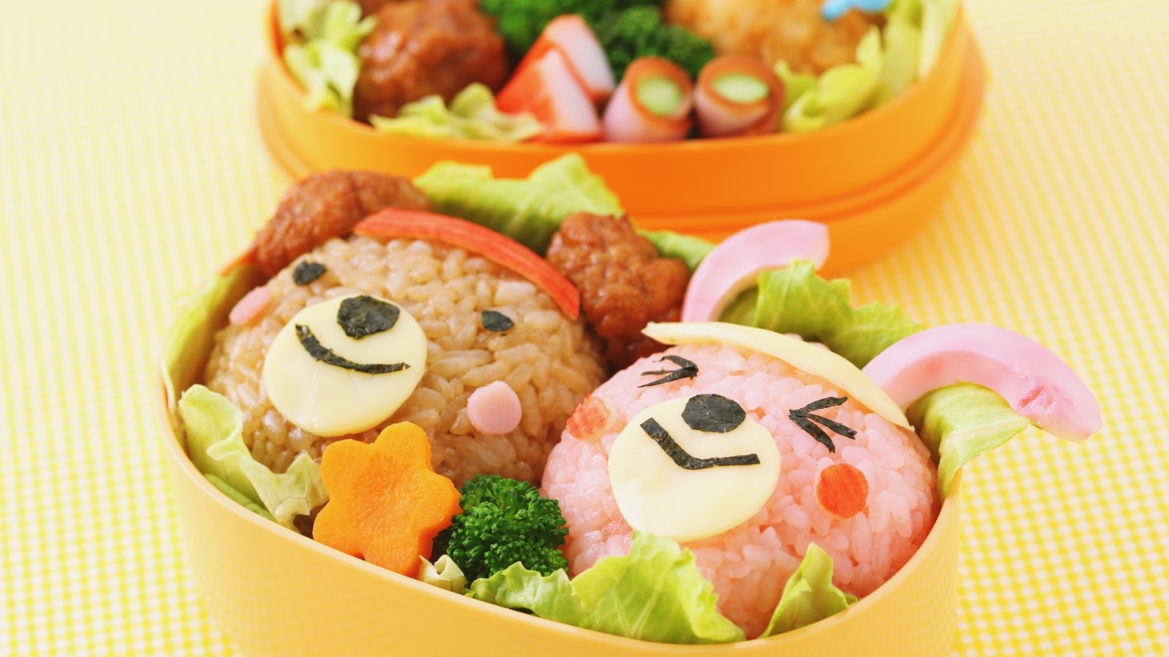 A colorful bento box features rice molded into a bear and bunny face, with cheese and seaweed details. Surrounded by fresh vegetables, it conveys a playful, cheerful tone.