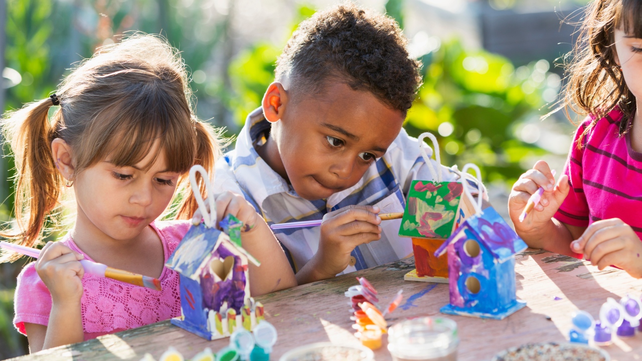 Children sit at an outdoor table, focused on painting colorful birdhouses. Bright, sunny setting with lush greenery in the background. Creative and joyful mood.