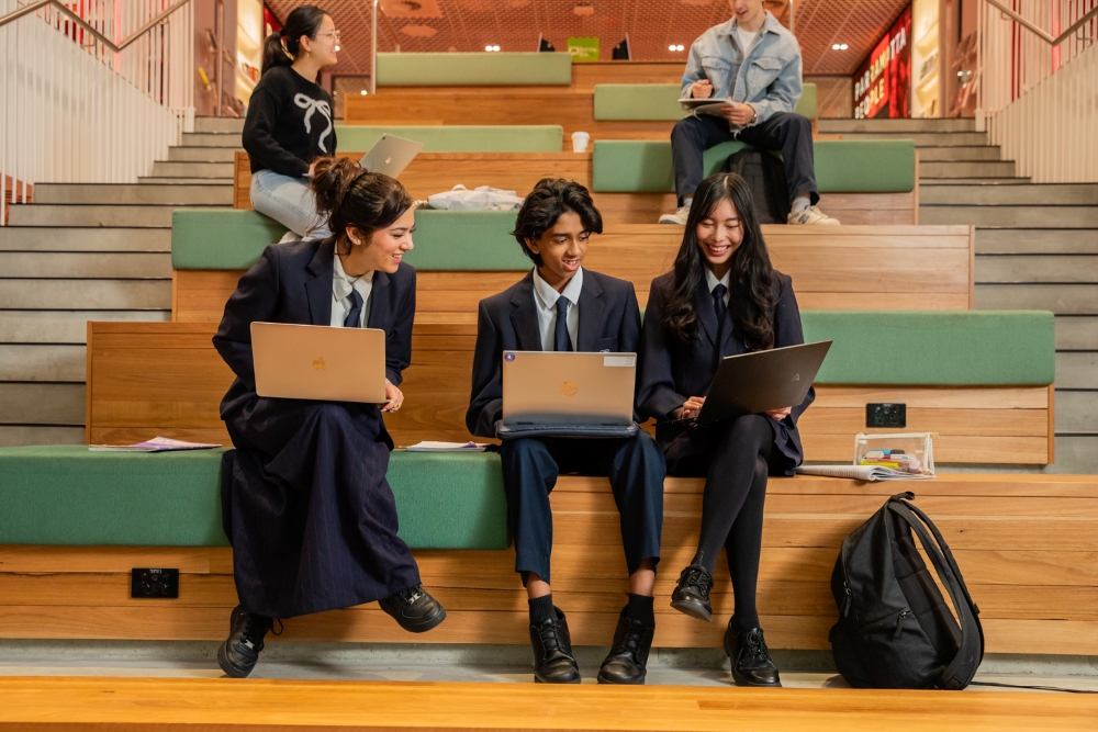 Three students in uniforms sit on wooden steps using laptops, smiling and collaborating. Two more students study in the background, creating a focused and lively atmosphere.