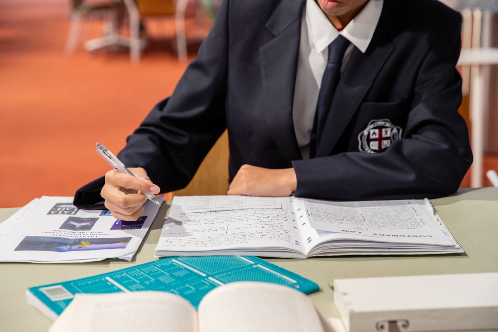 A person in a school uniform studies at a table, writing notes in an open binder. Books and papers are scattered, creating a focused atmosphere.