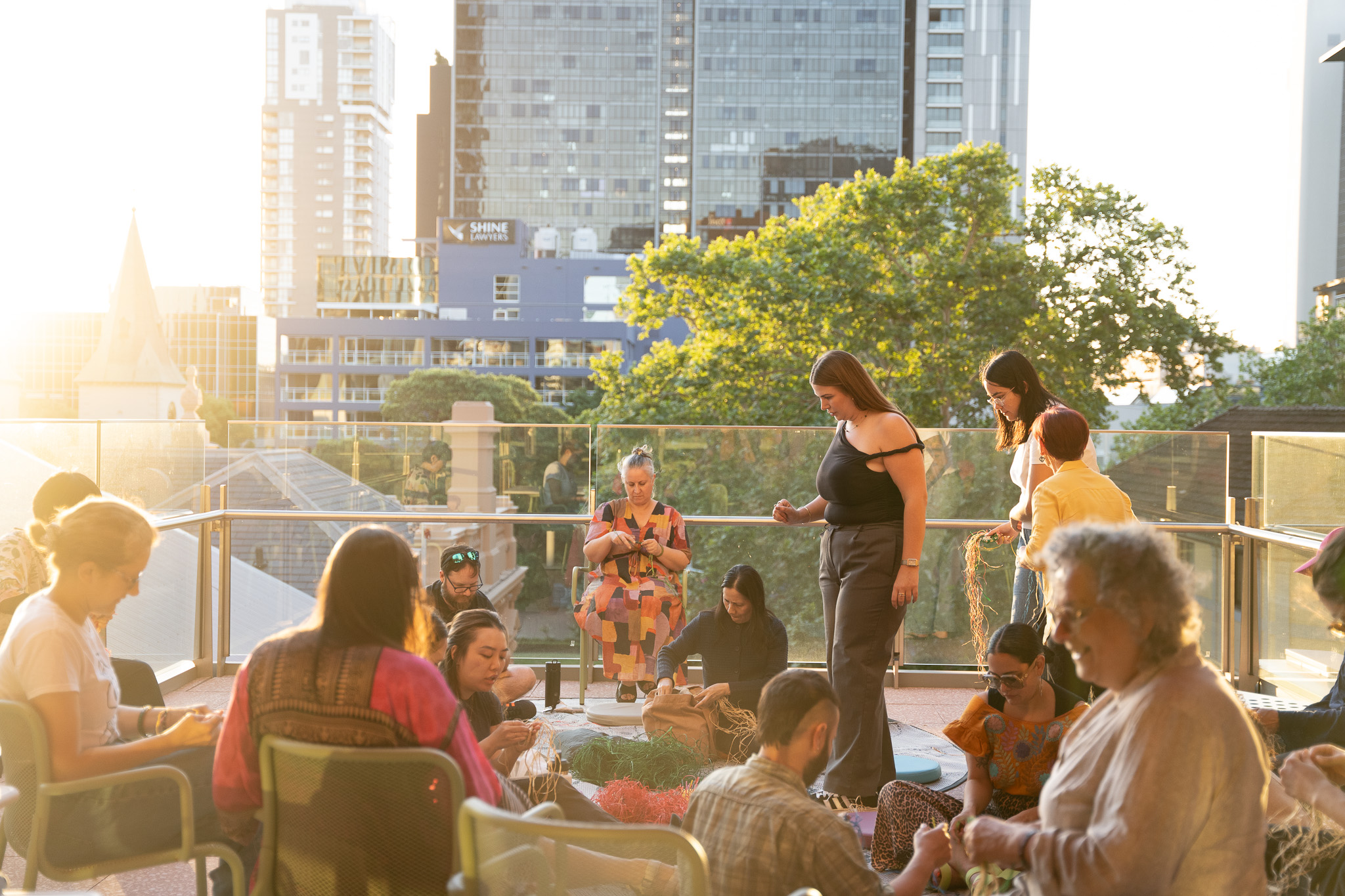 A group of people gathered on a sunlit terrace, engaging in a weaving activity. They are surrounded by urban buildings and trees, creating a warm, communal atmosphere.