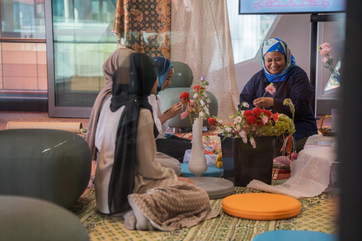 Four women in colorful headscarves sit on cushions, joyfully arranging flowers in a bright, cozy room. The scene exudes warmth and creativity.