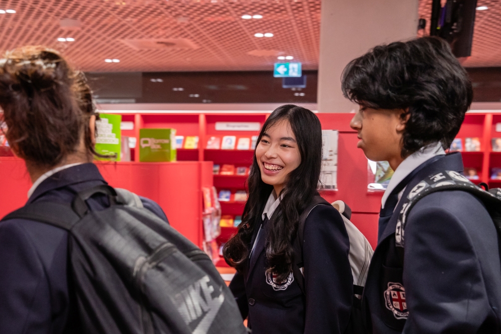 Three students in school uniforms converse and smile inside a library with red shelves filled with books. The mood is lively and engaging.