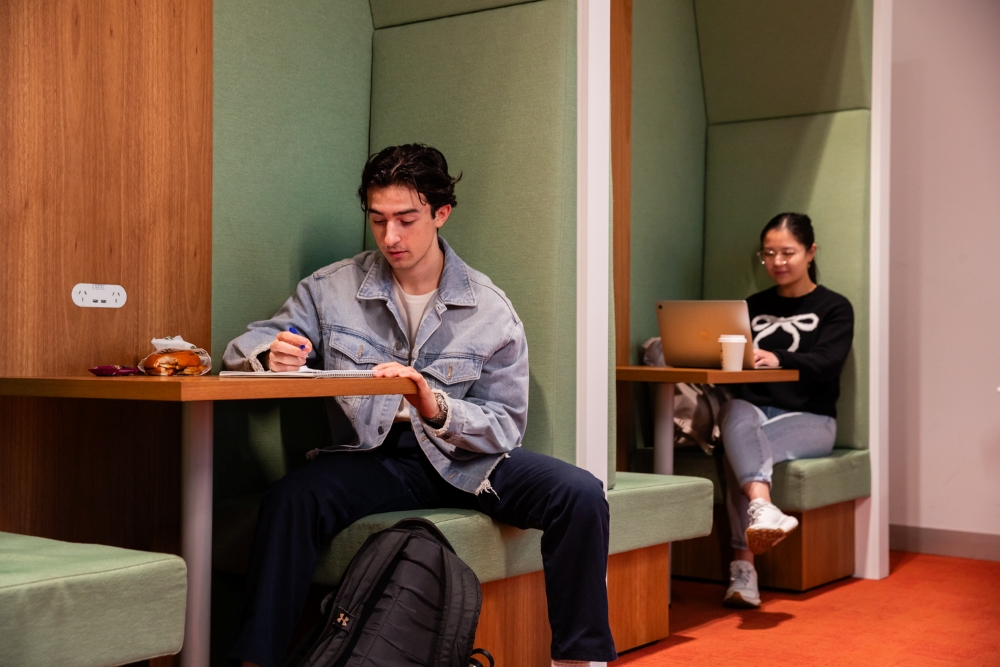 Two students study in separate booth seats. The man in a denim jacket writes in a notebook, while the woman uses a laptop with a coffee nearby. The atmosphere is focused.