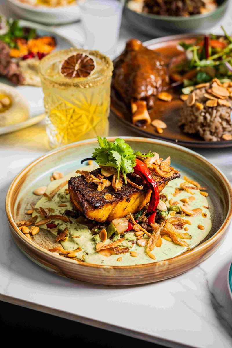 A vibrant table of food, focused on a fish dish, lamb shanks and rice and a colourful yellow cocktail.