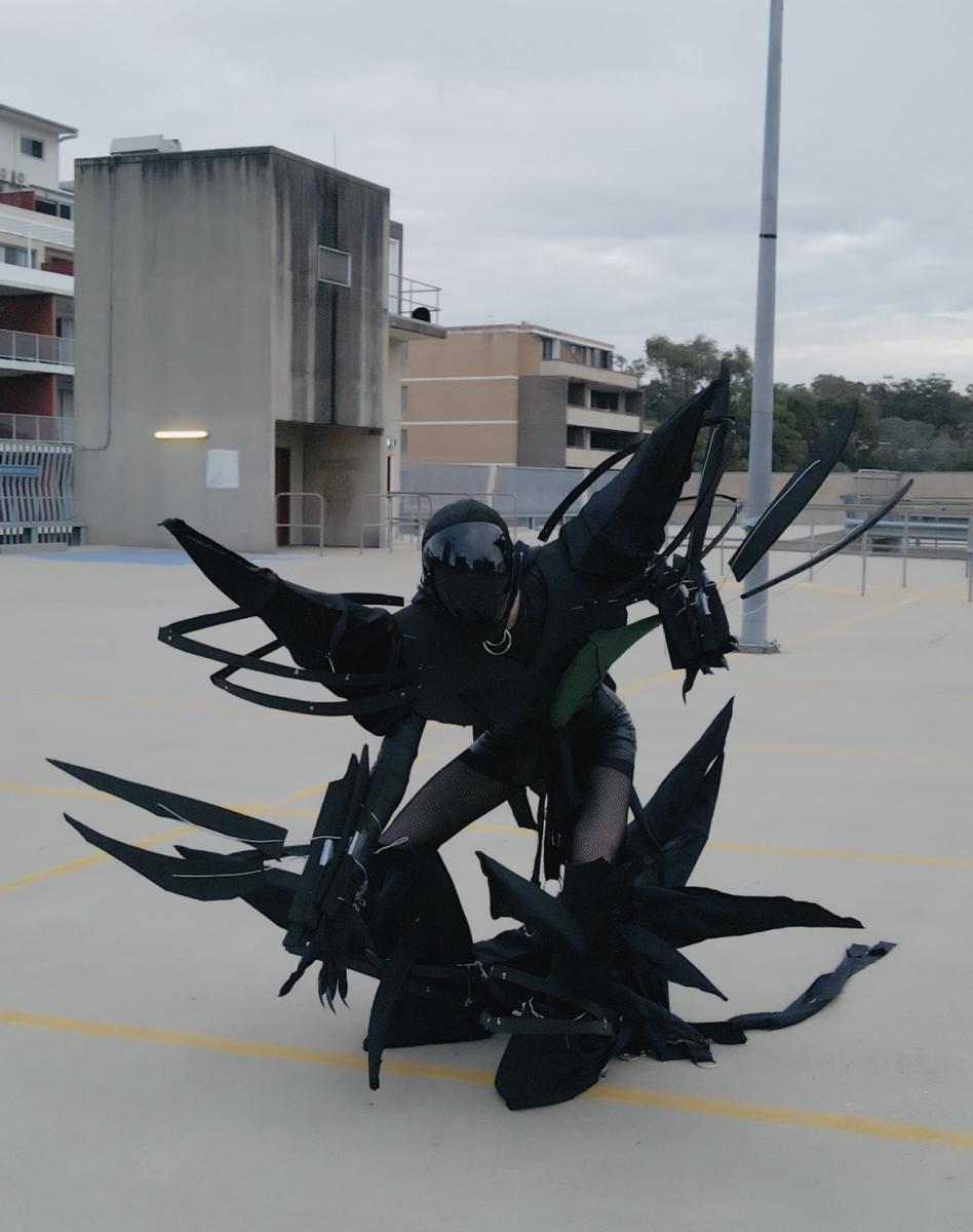 A black costumed figure stands along in a carpark