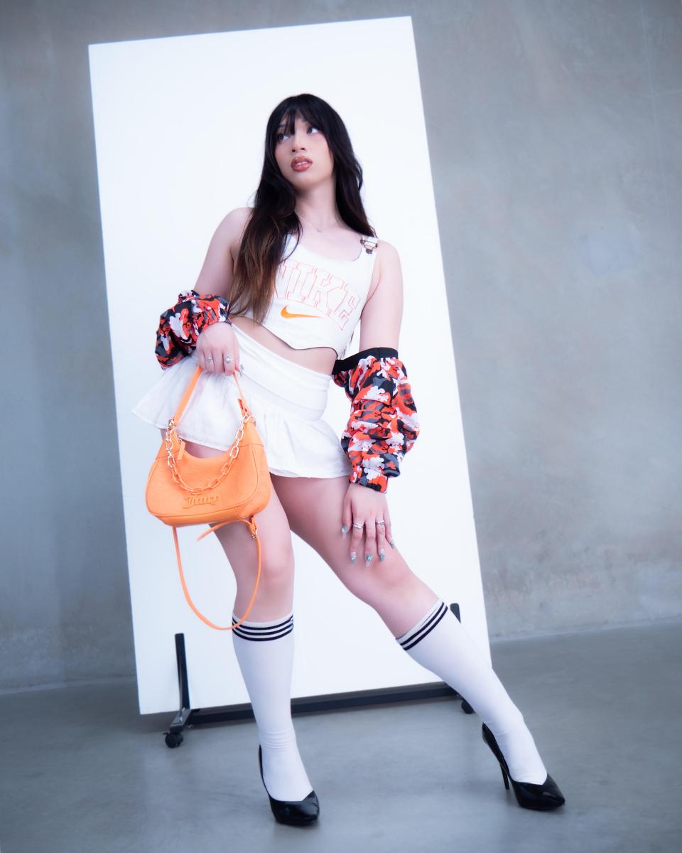 A woman in a sporty-chic outfit poses confidently. She wears a white crop top, mini skirt, long socks, heeled shoes, and holds an orange handbag, exuding a playful, stylish vibe.