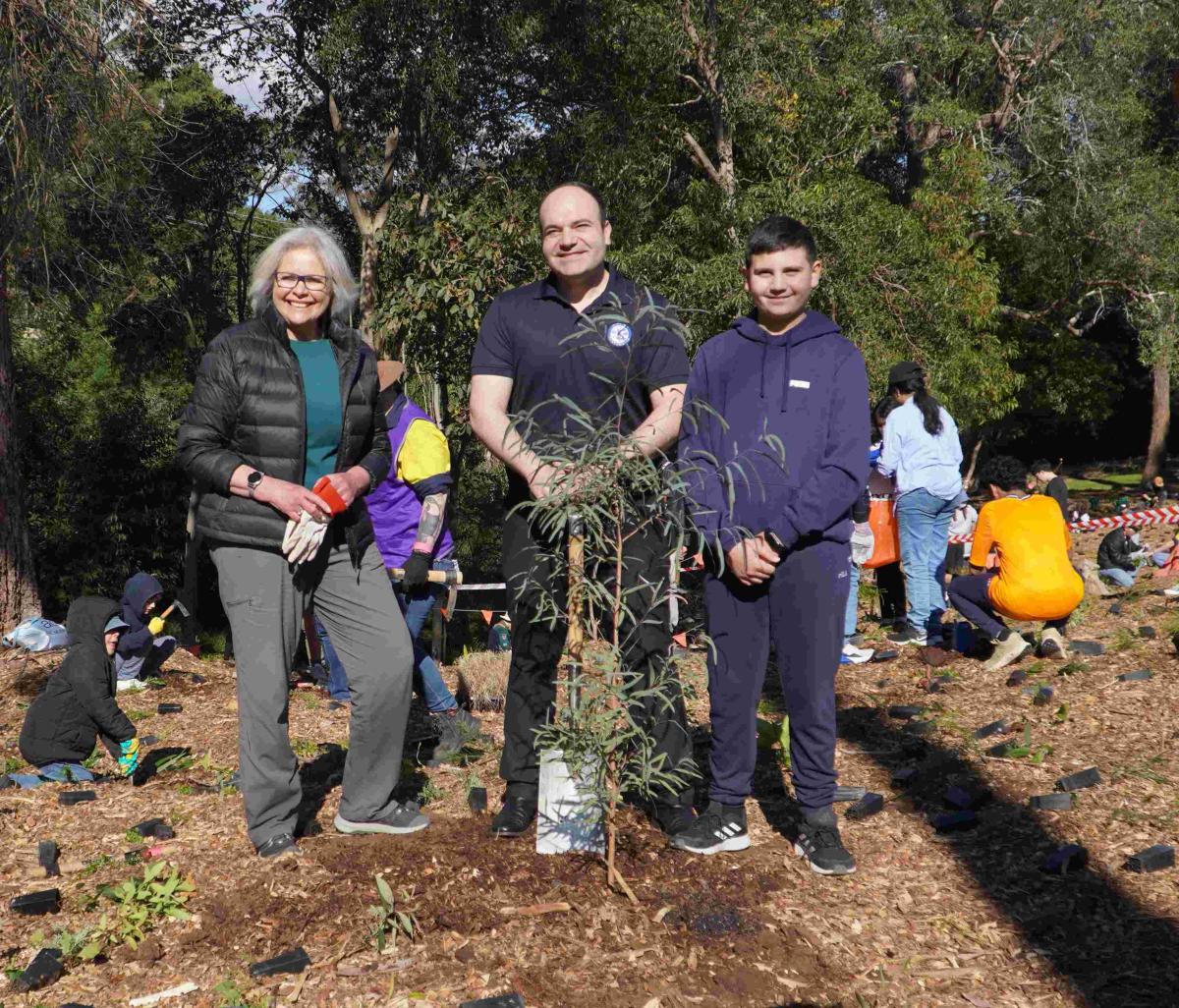 Three people stand smiling in a park during a tree planting event, surrounded by others planting saplings. The mood is community-focused and joyful.
