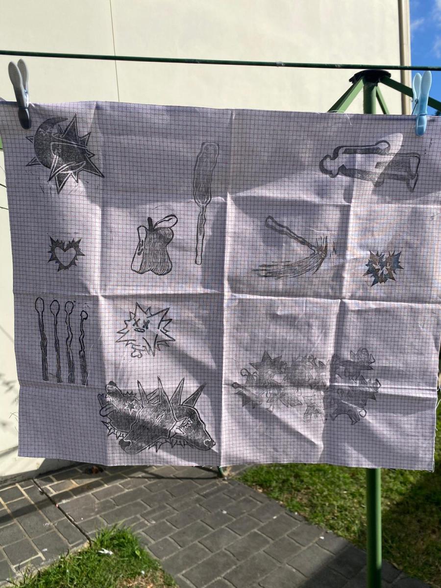 A graph paper towel with various hand-drawn symbols and abstract shapes is clipped to a clothesline under a blue sky. The scene is bright and casual.