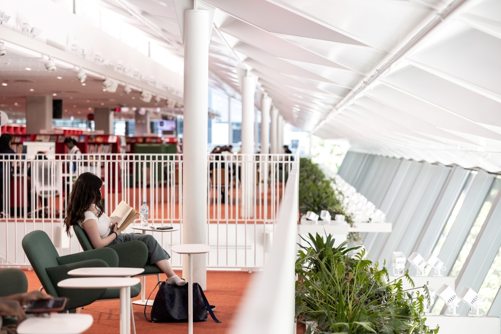 A woman reads a book in a modern library with red carpet and green plants. She sits on a green chair beside a white railing, creating a calm atmosphere.