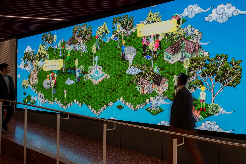 Large digital screen with vibrant pixel art depicting a green island with trees, houses, and characters. Two men walk by, wearing business attire.