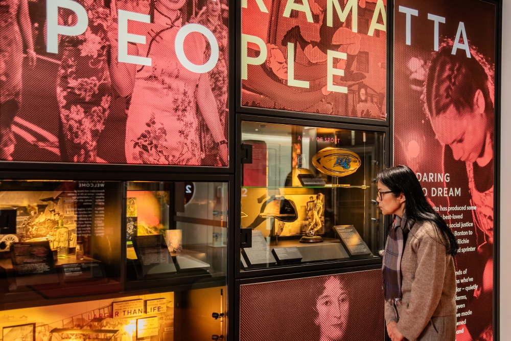 A person observes a museum exhibit featuring cultural artifacts and historical images with signage reading "PEOPLE." The scene is vibrant and contemplative.