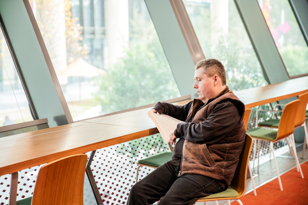A man sits by large windows in a modern library, engrossed in reading a book. The bright, airy space conveys a calm and focused atmosphere.