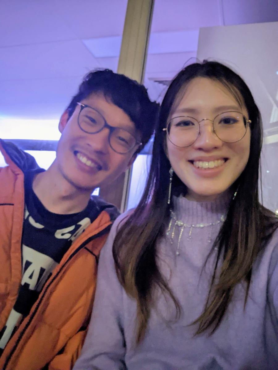 Two people with glasses smile brightly for a selfie. The person on the left wears an orange jacket, and the one on the right is in a lavender sweater. Warm, cheerful mood.