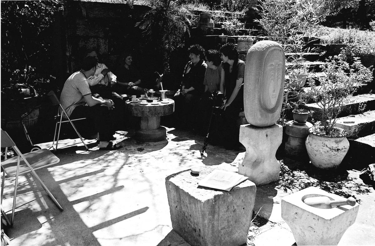 Black and white photo of a group of people seated in a sunny garden. They gather around a small table with cups, near sculptures and potted plants. The mood is relaxed.