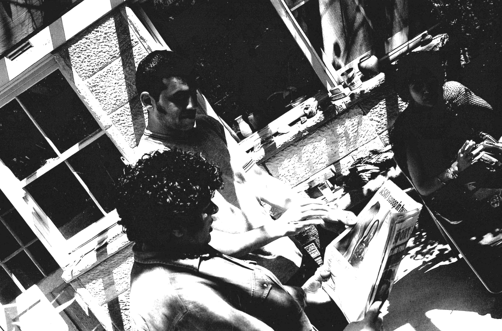 Three people gather around a table outdoors in high contrast black and white. One holds a magazine. Sunlight filters through leaves, casting shadows.