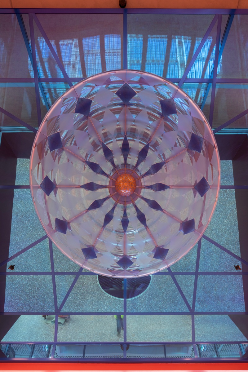 A colorful, geometric dome lights up with intricate patterns and a central orange hub. The reflection on blue-framed glass beneath creates a kaleidoscopic effect.