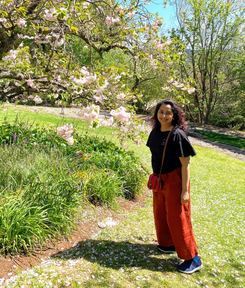 A woman stands smiling in a sunny garden, surrounded by blooming pink cherry blossoms and lush greenery. She's wearing a black top and red pants.