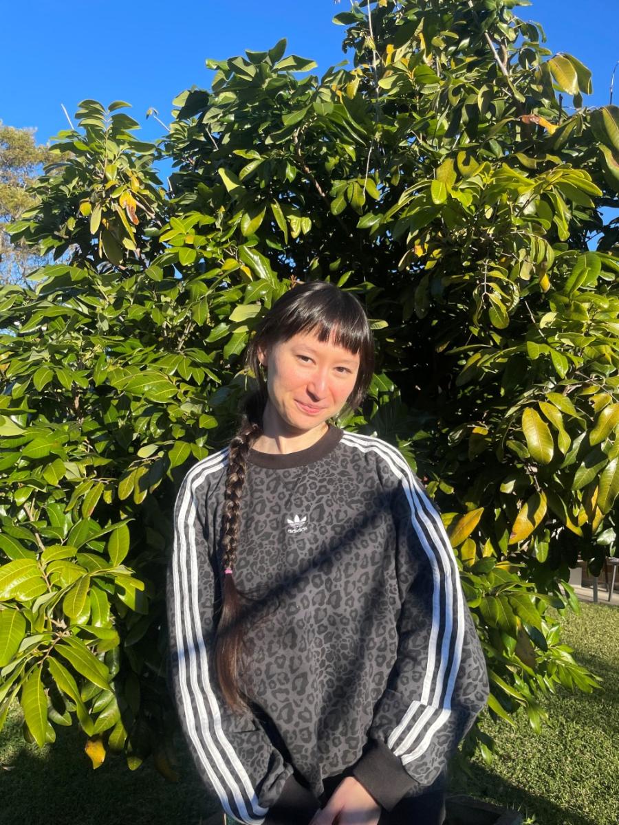 A person with long braided hair stands smiling in front of a lush, green tree under a clear blue sky, wearing a patterned Adidas sweatshirt.