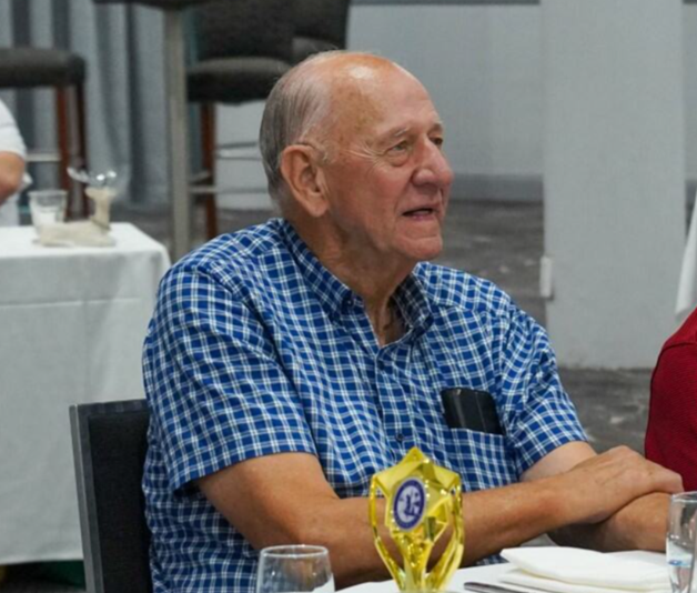 Elderly man in blue checked shirt sitting down at a table with an award in front of him