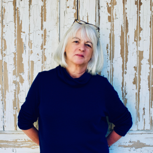 Author Tea Cooper wearing a blue coat standing in front of a timber white wall