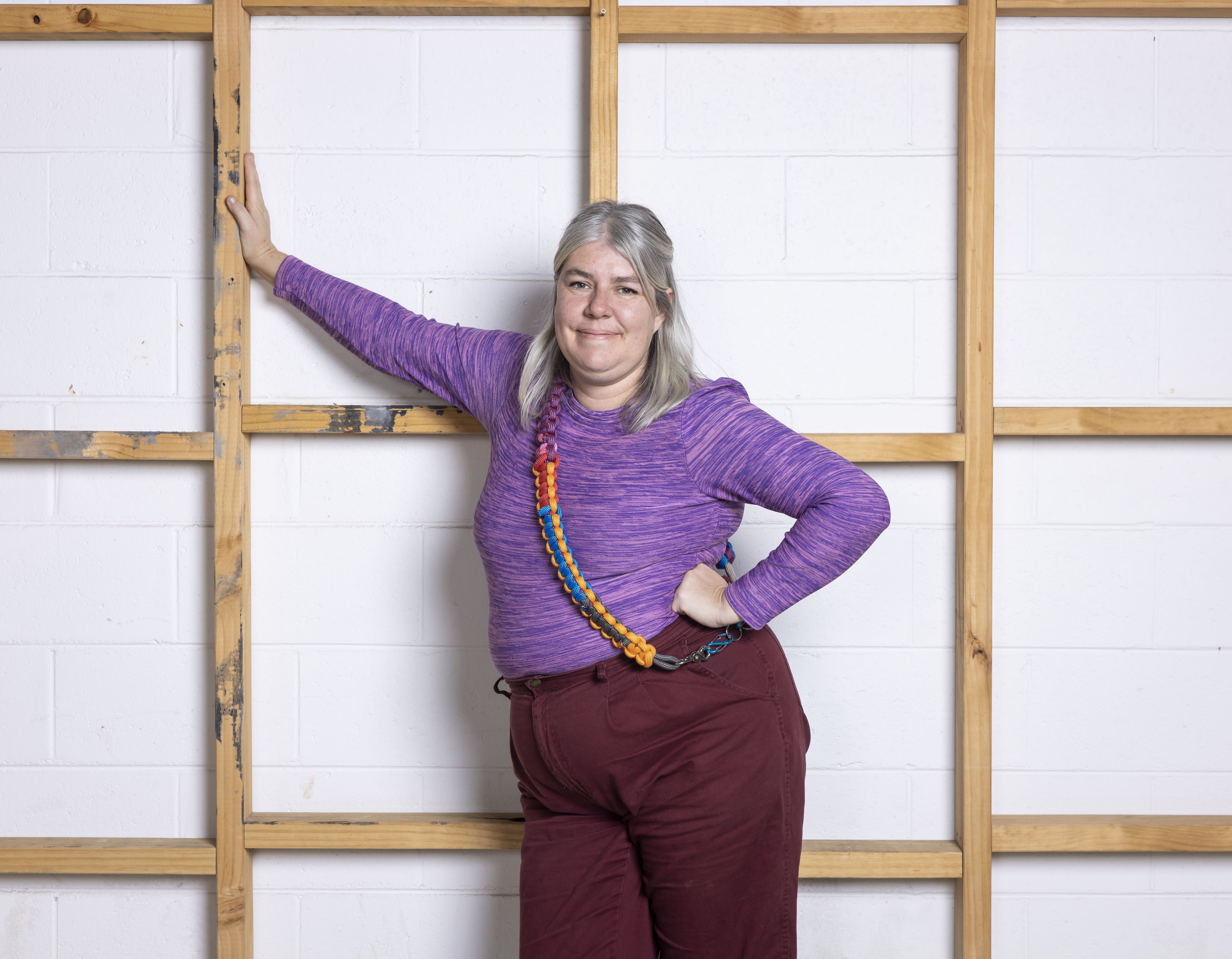 A woman with grey hair and brightly coloured clothing stands leaning against a wooden stud wall