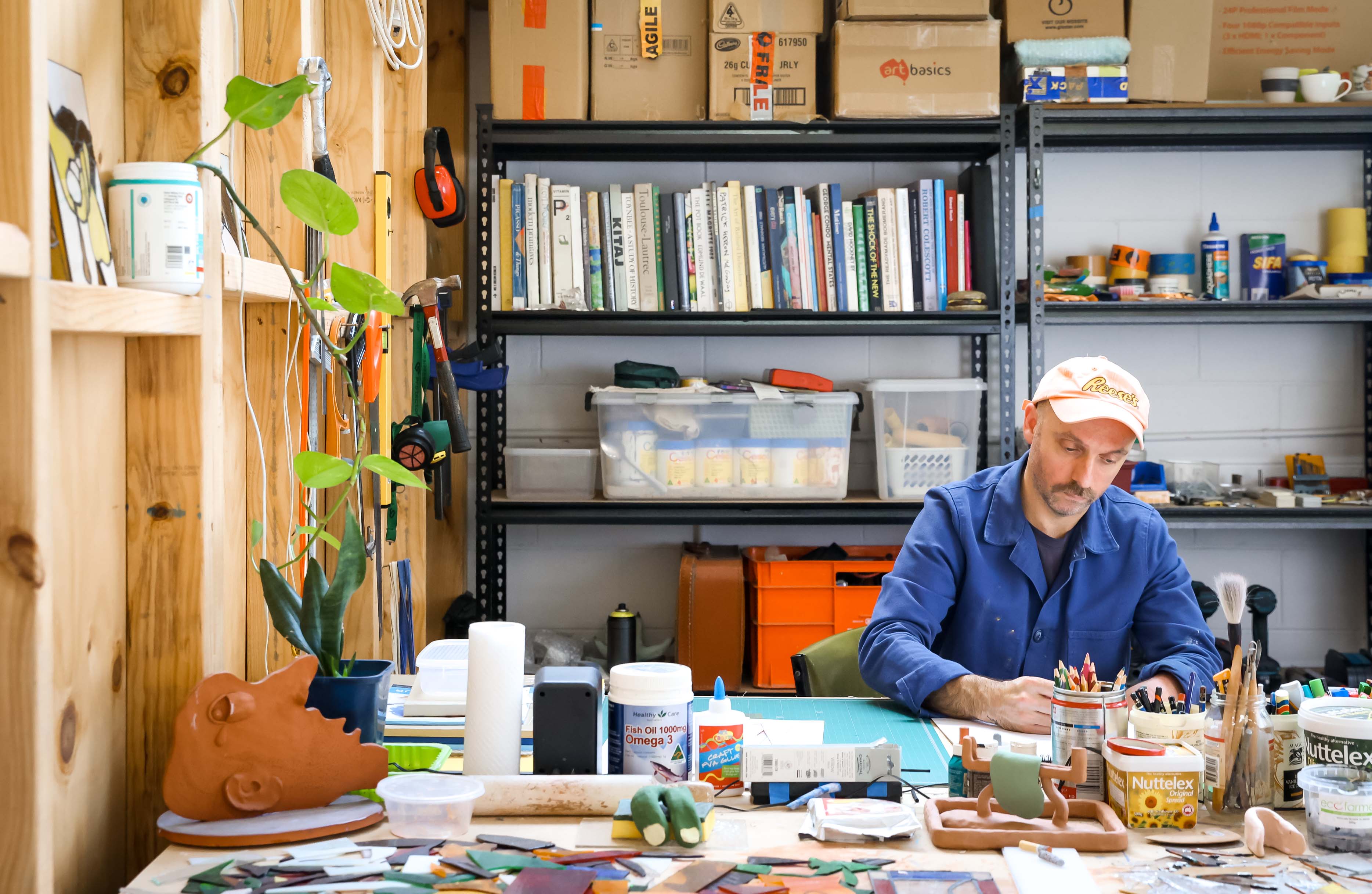 A man in a blue shirt and white cap works at a table filled with art studio materials