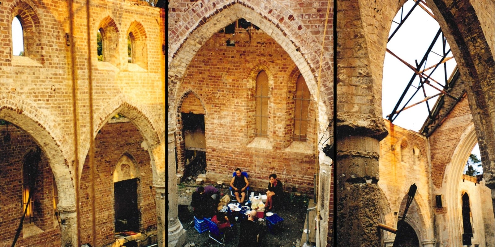 People sit around a table enjoying a meal inside a roofless, aged brick building with tall arches and windows, creating a cozy yet historic atmosphere.