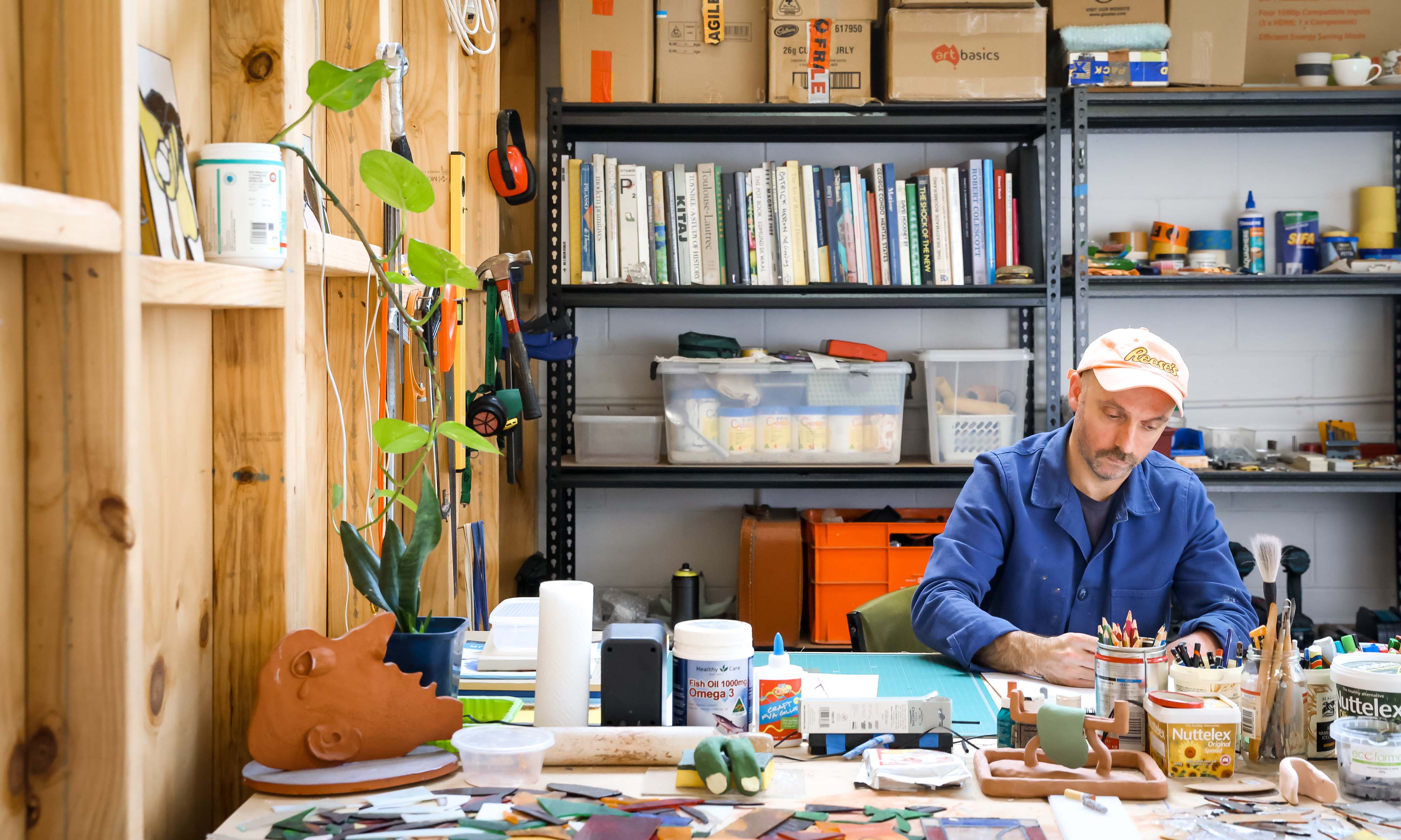 A man in a blue shirt and white cap sits in a studio filled with art and art materials