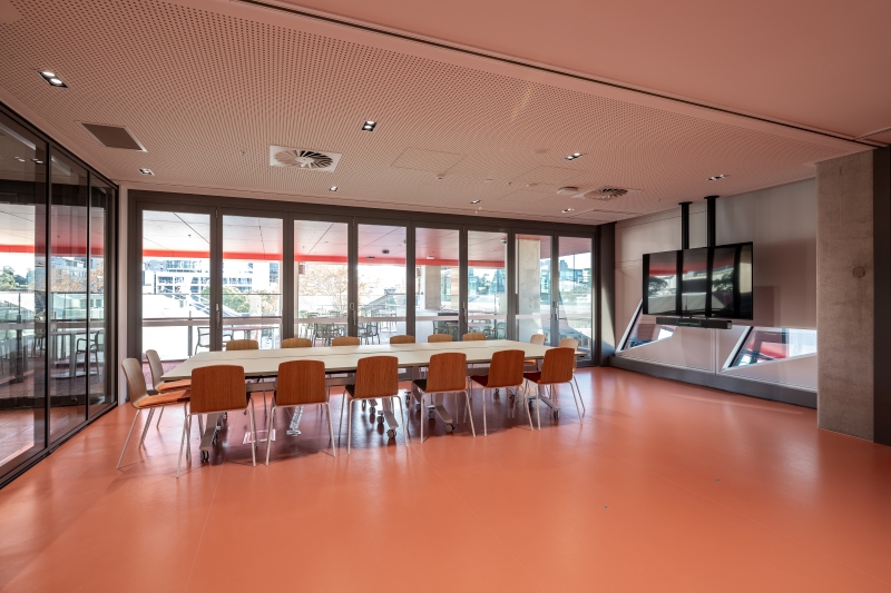 Modern conference room with a long white table, 10 wooden chairs, and a large TV on the wall. Glass walls reveal an outdoor patio. Bright, airy feel.
