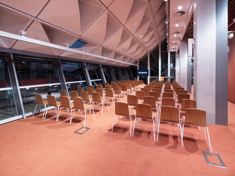 A modern conference room with rows of wooden chairs on an orange carpet. Large angled windows line one side, with a screen at the front. Calm atmosphere.