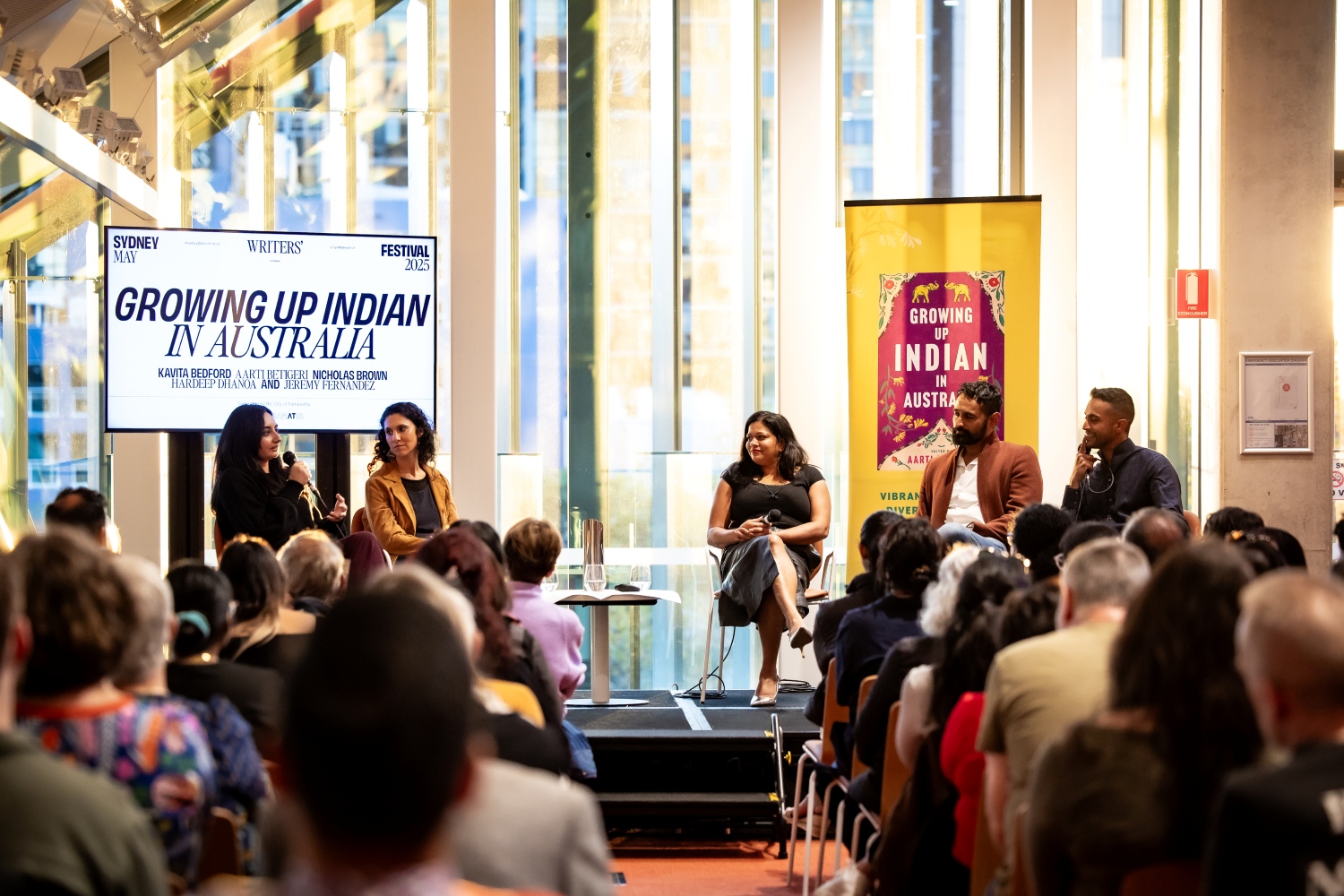 A panel of four speakers engage with an audience at the "Growing Up Indian in Australia" event during the Sydney Writers' Festival. They are seated on a stage with a large banner behind them displaying the event name. The atmosphere is attentive and focused.