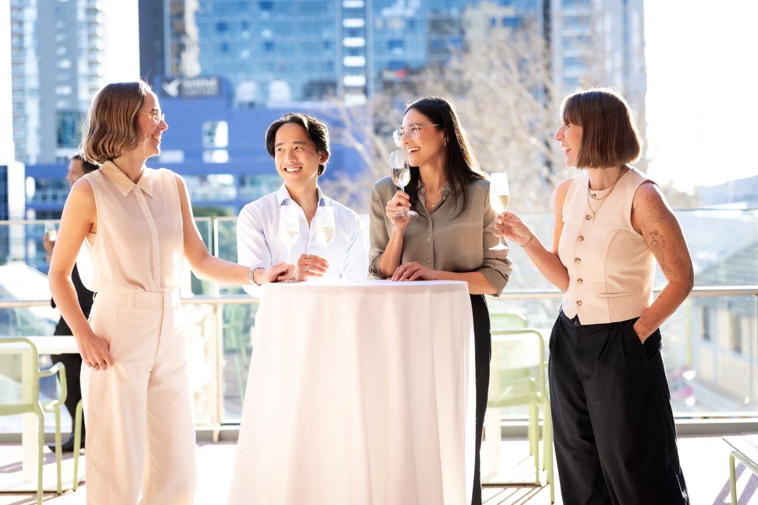 Four people stand around a table outdoors, smiling and holding wine glasses, with a cityscape in the background, conveying a joyful, social atmosphere.