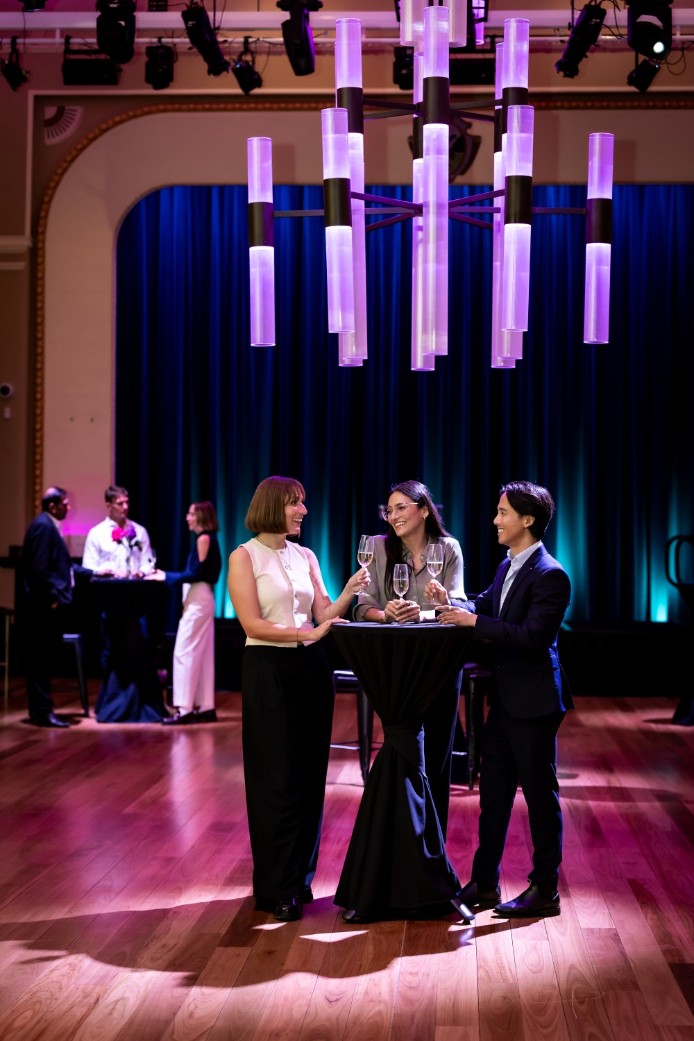 Three people in formal attire enjoy a toast at a cocktail table under purple lighting in an elegant venue, creating a celebratory atmosphere.